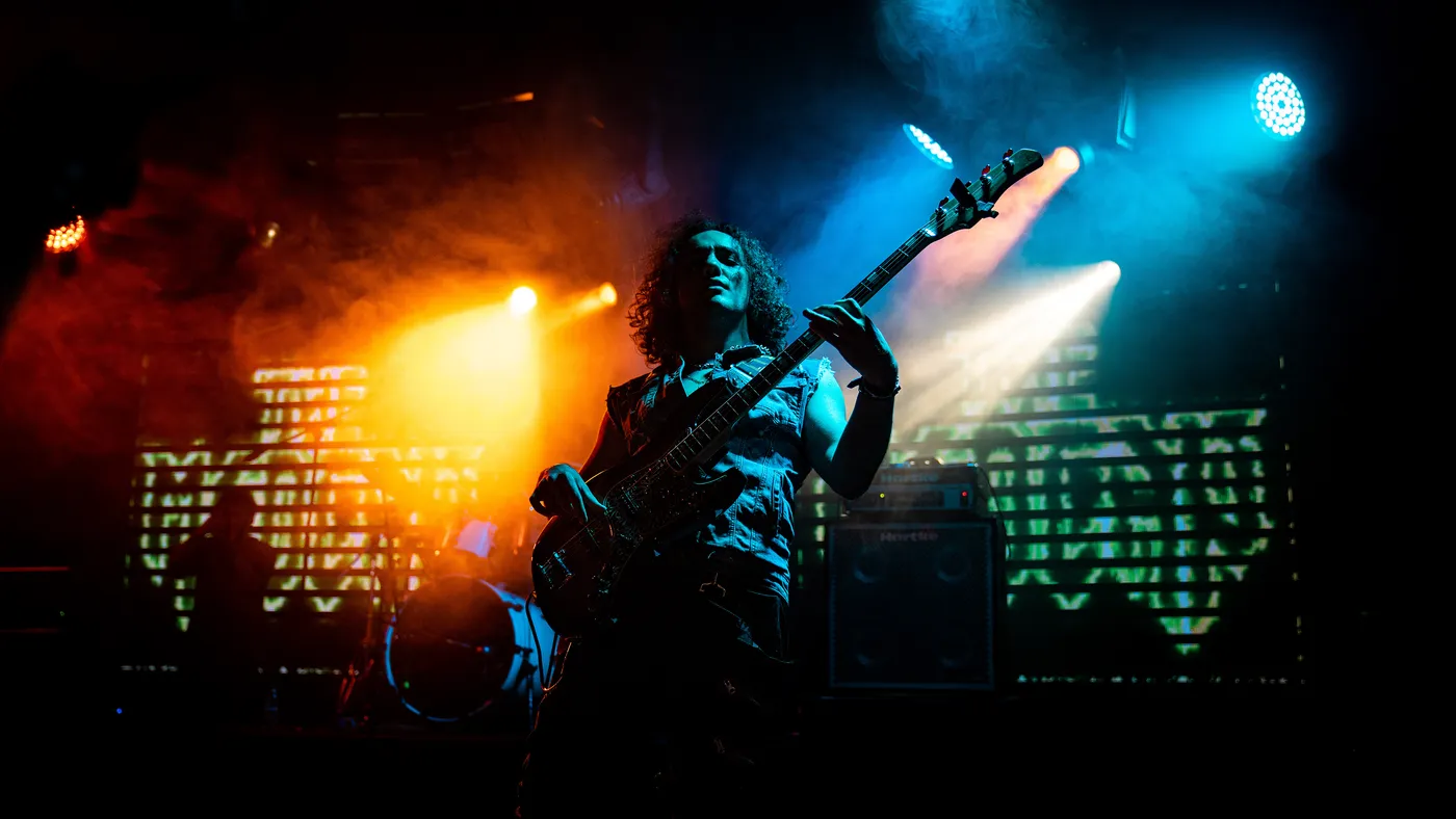Konser sahnesinde arkadan gelen turuncu ve mavi ışıklar arasında gitar çalan müzisyen